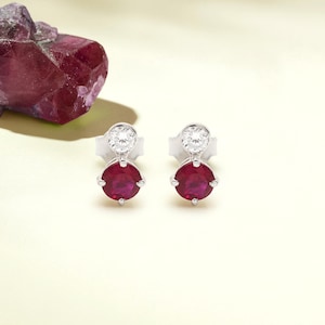 May include: Silver stud earrings with a round, deep red ruby and a smaller, clear gemstone. The earrings are set in silver. A rough, dark red gemstone is in the background. The earrings are a classic jewellery design.