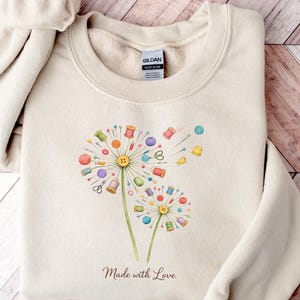 May include: Beige sweatshirt featuring a dandelion design with colorful sewing notions as seeds. The dandelion has spools of thread, scissors, and buttons. The text "Made with Love" is printed below the design. The sweatshirt is folded on a wooden surface.