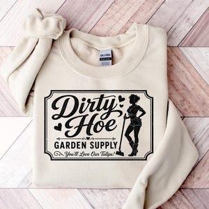 Puede incluir: Una sudadera color crema con cuello redondo. La parte delantera presenta un gráfico negro que dice "Dirty Hoe Garden Supply" con la silueta de una mujer sosteniendo una herramienta de jardinería. También se incluye el texto "You'll Love Our Tulips!".