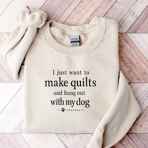 Puede incluir: Una sudadera de cuello redondo color arena con el texto impreso en negro "I just want to make quilts and hang out with my dog" seguido de un pequeño icono de huella y guiones decorativos. La prenda tiene una etiqueta de la marca Gildan.