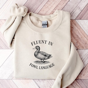 May include: A cream-colored crewneck sweatshirt featuring a black graphic of a duck in the center. The text "FLUENT IN" is printed above the duck, and "FOWL LANGUAGE" is printed below the duck in a black serif font. The garment has a Gildan label.