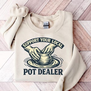 May include: A beige sweatshirt with the text "SUPPORT YOUR LOCAL POT DEALER" in a black and cream graphic. The graphic depicts hands shaping a pot on a pottery wheel. The sweatshirt is folded on a wooden surface.