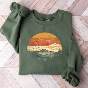 May include: A green sweatshirt with a circular graphic depicting a mountain scene at sunset. The design includes a yellow and orange sun, mountains, trees, and birds. The sweatshirt has a crew neck and long sleeves.