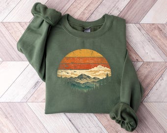 May include: A green sweatshirt featuring a circular graphic of a mountain landscape with a sunset design. The graphic includes a yellow and orange sun, mountains, trees, and birds. The sweatshirt has a crew neck and long sleeves.