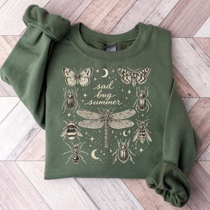 May include: An olive green crewneck sweatshirt featuring a cream-colored graphic print of various insects including a dragonfly, beetles, a bee, and butterflies, surrounding the text "sad bug summer" with small stars and crescent moon accents.