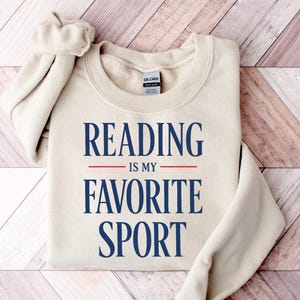 May include: A cream-colored sweatshirt with the text "READING IS MY FAVORITE SPORT" in navy blue. The text is centered, with a red line separating "READING" and "IS MY". The sweatshirt is folded on a wooden surface.