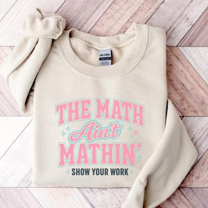 May include: A sand-colored crewneck sweatshirt featuring the text THE MATH AIN'T MATHIN' in pink and light blue block lettering, with the phrase SHOW YOUR WORK in dark gray below, accented by small light blue and pink star-shaped sparkles.