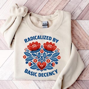 Könnte beinhalten: Ein cremefarbenes Sweatshirt mit dem Text "RADICALIZED BY BASIC DECENCY" in Blau. Das Design zeigt ein florales Herz in Rot und Blau. Das Sweatshirt liegt auf einer Holzoberfläche.