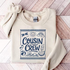 May include: A cream-colored crewneck sweatshirt featuring a dark blue graphic print of a beach towel border containing the text "COUSIN CREW" surrounded by illustrations of sunglasses, a sun, a rainbow, strawberries, a beach ball, waves, and a popsicle.