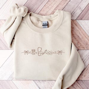 May include: A sand-colored crewneck sweatshirt featuring a brown line-art embroidery across the chest. The design includes a bow, a pill bottle, a stethoscope, a syringe, a bandage, and another bow, all connected by a continuous flowing line.