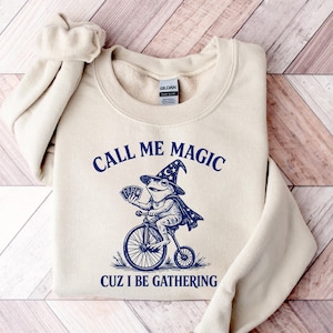 Op de afbeelding: Een beige sweater met ronde hals en een donkerblauwe grafische print van een kikker met een tovenaarshoed en cape, rijdend op een hoge bi terwijl hij speelkaarten vasthoudt. De tekst luidt CALL ME MAGIC CUZ I BE GATHERING in blauwe inkt.