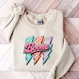 May include: A cream-colored crewneck sweatshirt featuring a graphic of three lightning bolts with a checkered pattern in teal, coral, and lavender. The word Boone is printed in a bold, pink, cursive font with a black outline across the bolts.