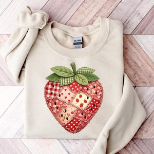 May include: A cream-colored sweatshirt featuring a large, patchwork strawberry design. The strawberry is composed of various red and pink patterned fabrics, including polka dots, gingham, and floral prints, with green leaves and stem.