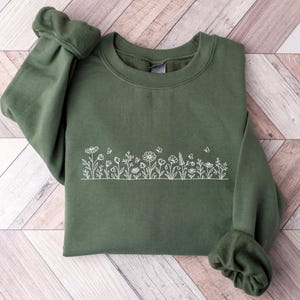 May include: An olive green crewneck sweatshirt featuring a white line art embroidery of a wildflower meadow across the center. The design includes various stems, leaves, small blossoms, and tiny butterflies arranged in a horizontal row.