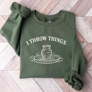 May include: An olive green crewneck sweatshirt featuring a white graphic print on the chest. The design includes the text I THROW THINGS arched above a line drawing of a ceramic vase centered on a pottery wheel, with small circular accents.