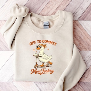 May include: A beige crewneck sweatshirt featuring a graphic of a white duck wearing heart-shaped sunglasses and a bandana while riding a scooter. The text above the duck reads OFF TO COMMIT and the text below reads Mom Foolery in orange.