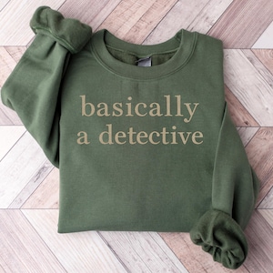 May include: An olive green sweatshirt with the phrase "basically a detective" printed in a light tan color. The sweatshirt is folded, with the sleeves rolled up. The background is a light wood pattern.