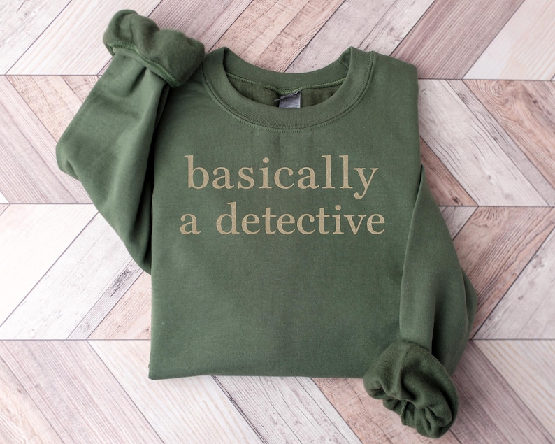 May include: An olive green sweatshirt with the phrase "basically a detective" printed in a light tan color. The sweatshirt is folded, with the sleeves rolled up. The background is a light wood pattern.
