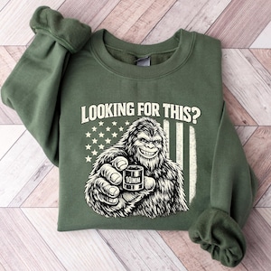 Puede incluir: Sudadera verde con cuello redondo con un gráfico de Bigfoot sonriendo sosteniendo un cartucho de 10 mm, sobre un diseño de bandera estadounidense desgastada. El texto "LOOKING FOR THIS?" está encima del gráfico. La sudadera tiene mangas enrolladas.
