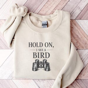 May include: A sand-colored crewneck sweatshirt featuring the dark gray text "HOLD ON, I SEE A BIRD" printed above a detailed black and white illustration of a pair of binoculars. The garment has long sleeves and a ribbed collar.