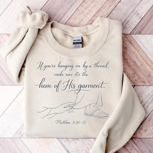 May include: A cream-coloured sweatshirt featuring the quote "If you're hanging on by a thread, make sure it's the hem of His garment." and a line drawing of a hand touching a foot. The text "Matthew 9:20-22" is also included.
