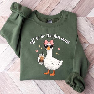 May include: Green sweatshirt featuring the text "off to be the fun aunt" and a goose graphic wearing sunglasses and a bow. The goose is holding a drink with a straw. The design also includes pink hearts and sparkles.
