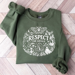 Può includere: Una felpa girocollo verde oliva con una stampa grafica bianca che recita "RESPECT THE LOCALS" circondata da illustrazioni di una falena, un sasquatch, un alieno, un serpente marino, fantasmi, funghi, alberi e un teschio di cervo.