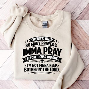 以下が含まれることがあります： 黒い文字で「THERE'S ONLY SO MANY PRAYERS IMMA PRAY BEFORE I CUSS YOU OUT. + I'M NOT FINNA KEEP BOTHERIN' THE LORD.」と書かれたベージュのスウェットシャツ。袖は折り返されています。