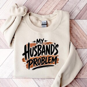 May include: A beige crewneck sweatshirt with the phrase "MY HUSBAND'S PROBLEM" in black and brown lettering. The sweatshirt is folded on a wooden surface.