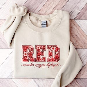 Puede incluir: Una sudadera de cuello redondo color arena que presenta la palabra RED en letras grandes de bloque rellenas con un patrón floral rojo y rosa. Debajo de la palabra, el texto remember everyone deployed está impreso en una fuente cursiva roja.