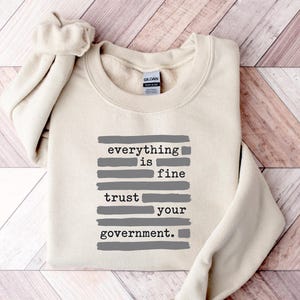 Può includere: Una felpa girocollo color crema con un design grafico composto da pennellate orizzontali grigie e dal testo nero everything is fine trust your government. Il capo presenta maniche lunghe e un'etichetta del marchio Gildan sul colletto.