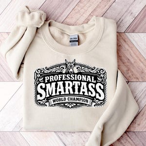Puede incluir: Una sudadera beige con el texto "PROFESSIONAL SMARTASS WORLD CHAMPION" en un diseño decorativo en blanco y negro. El diseño incluye una ilustración de un burro. La sudadera está doblada sobre una superficie de madera.