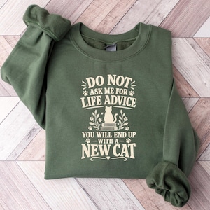 May include: An olive green crewneck sweatshirt featuring cream-coloured text that reads DO NOT ASK ME FOR LIFE ADVICE YOU WILL END UP WITH A NEW CAT. The design includes a graphic of a cat sitting on a stack of books surrounded by floral accents.