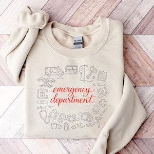 May include: A sand-colored crewneck sweatshirt featuring a central graphic with the red text "emergency department" surrounded by black line-art illustrations of medical equipment including an ambulance, stethoscope, syringe, and heart monitor.