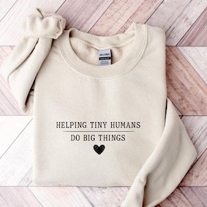 May include: A beige crewneck sweatshirt featuring the black printed text HELPING TINY HUMANS DO BIG THINGS with a small black heart icon centred below the text. The garment has long sleeves and a ribbed collar with a Gildan brand label inside.