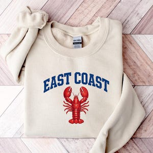 Puede incluir: Una sudadera de cuello redondo color crema con el texto EAST COAST en letras azules arqueadas sobre una ilustración detallada de una langosta roja brillante centrada en el pecho. La prenda cuenta con mangas largas y cuello acanalado.