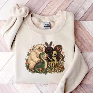 Può includere: Una felpa girocollo color crema con una stampa grafica raffigurante una creatura bianca pelosa, un rettile acquatico verde, una falena nera con ali macchiate di arancione e un piccolo alieno verde seduti insieme tra i funghi.