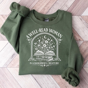 May include: An olive green crewneck sweatshirt featuring a white graphic print of an open book with flowers and butterflies emerging from the pages, framed by an arch. The text reads A WELL-READ WOMAN IS A DANGEROUS CREATURE in a serif font.