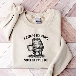 Puede incluir: Una sudadera de cuello redondo color arena con una ilustración gráfica en negro de un sapo sosteniendo una taza humeante. El texto sobre el sapo dice I HAVE TO SAY WEIRD y el texto debajo dice STUFF OR I WILL DIE en fuente negra.