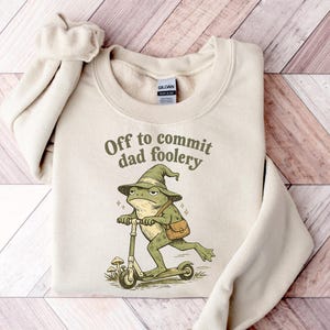 Puede incluir: Una sudadera color crema con la frase "Off to commit dad foolery" impresa en marrón. El gráfico presenta una rana verde con un sombrero de mago montada en un patinete. La rana tiene una bolsa marrón.