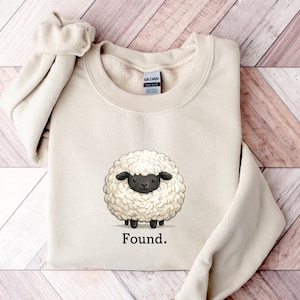 Könnte beinhalten: Ein beigefarbenes Sweatshirt mit Rundhalsausschnitt und einer Grafik eines flauschigen weißen Schafs mit schwarzem Gesicht und Beinen. Unter dem Schaf ist das Wort "Found." in einer einfachen schwarzen Serifenschrift gedruckt. Am Innenkragen befindet sich ein Gildan-Etikett.