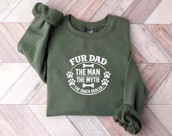 Peut inclure: Un sweat-shirt à col rond vert olive avec une inscription blanche imprimée FUR DAD THE MAN THE MYTH THE SNACK DEALER, entourée d'un cercle orné d'empreintes de pattes et d'icônes d'os, centré sur la poitrine du vêtement.