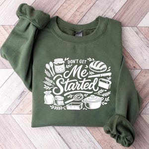 Op de afbeelding: Een olijfgroene sweater met ronde hals en een witte grafische print van keukengerei en bakbenodigdheden, waaronder een pot, een brood, een garde en een pan, met de tekst "DON'T GET ME STARTED" in een gestileerd handschrift.