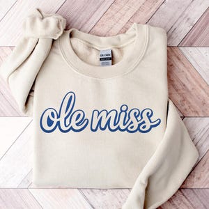 May include: A cream-colored sweatshirt with the words "Ole Miss" in a blue script font. The sweatshirt is folded on a wooden surface. The Gildan brand tag is visible on the collar.
