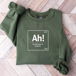 Op de afbeelding: Een olijfgroene sweater met ronde hals en een witte grafische print in de stijl van een element uit het periodiek systeem. De tekst luidt Ah! The Element of Surprise, met de getallen 29,7 en 230 in de rechterboven- en rechterbenedenhoek.