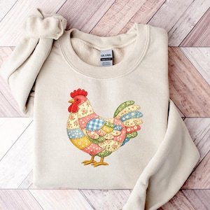 May include: A cream-coloured crewneck sweatshirt featuring a central graphic of a rooster composed of various colourful patchwork patterns, including floral, gingham, and polka dot designs in shades of red, yellow, blue, and green.
