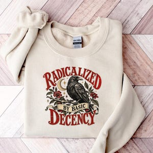 Puede incluir: Una sudadera de cuello redondo color crema con un estampado gráfico de un cuervo negro posado en una rama con una luna creciente y flores rojas. El texto dice RADICALIZED BY BASIC DECENCY en una fuente roja con remates.
