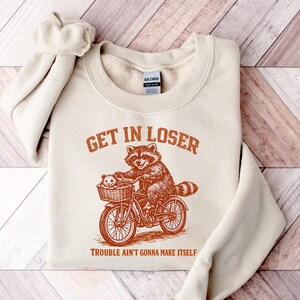 May include: A cream-colored crewneck sweatshirt featuring a rust-colored graphic of a raccoon riding a bicycle with an opossum in the front basket. The text reads GET IN LOSER above the raccoon and TROUBLE AIN'T GONNA MAKE ITSELF below.
