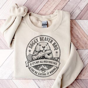 Może przedstawiać: Kremowa bluza z okrągłą grafiką. Grafika przedstawia bobra trzymającego kłodę i napis "Soggy Beaver BBQ". Zawiera również tekst "If it's not all over your face, you're eating it wrong". Bluza ma okrągły dekolt i długie rękawy.