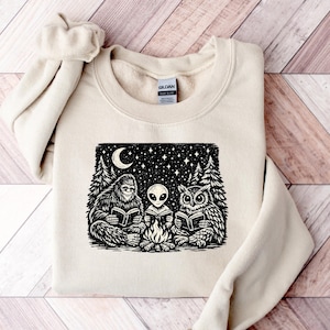 May include: A beige sweatshirt featuring a black and white graphic of Bigfoot, an alien, and an owl reading books around a campfire under a starry night sky. The design includes a crescent moon and pine trees.
